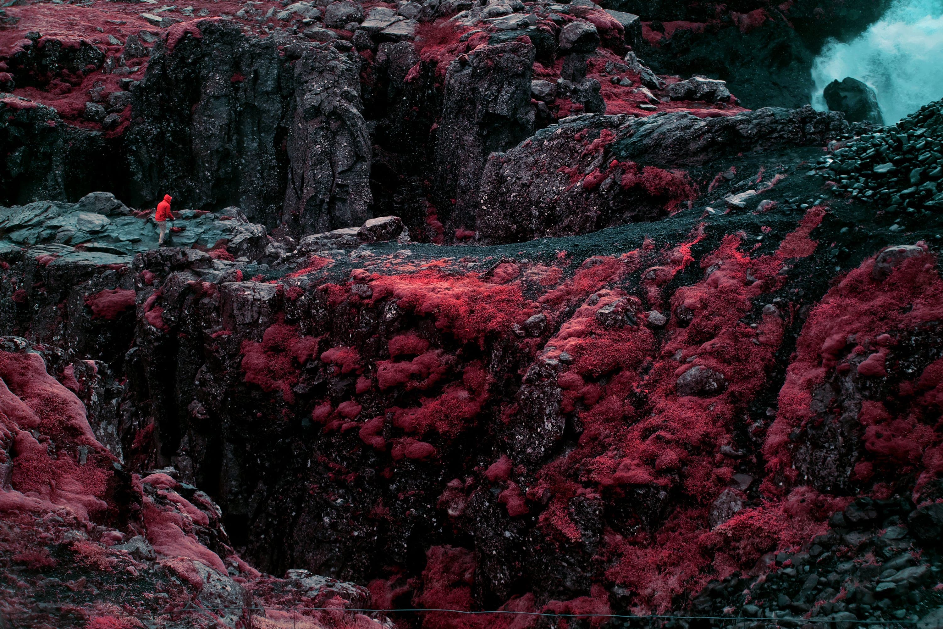A distant figure in red standing at a dark cliff edge above slopes of ember-red moss