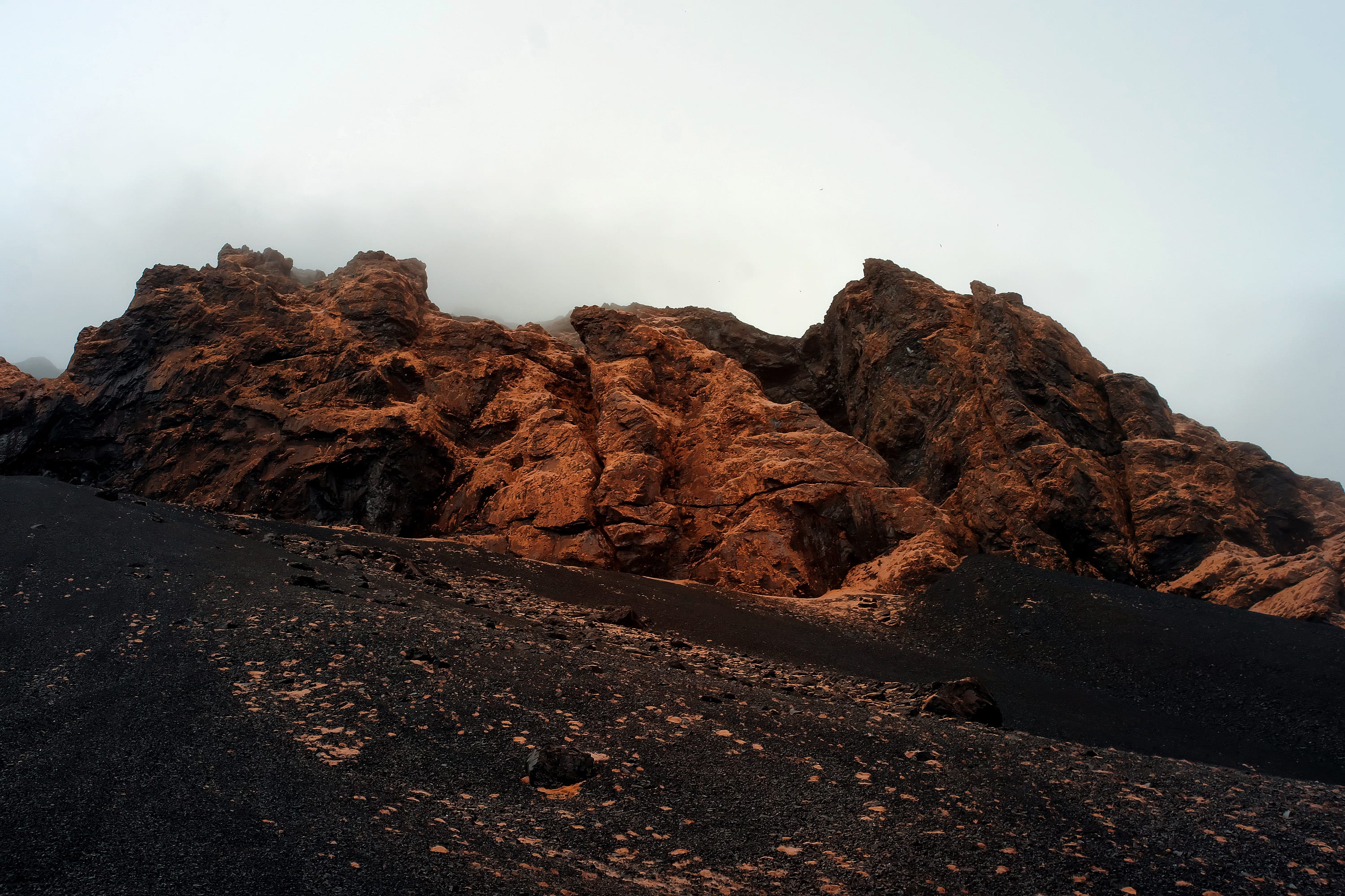 Rust-orange rock outcrops above a black ash slope dissolving into fog