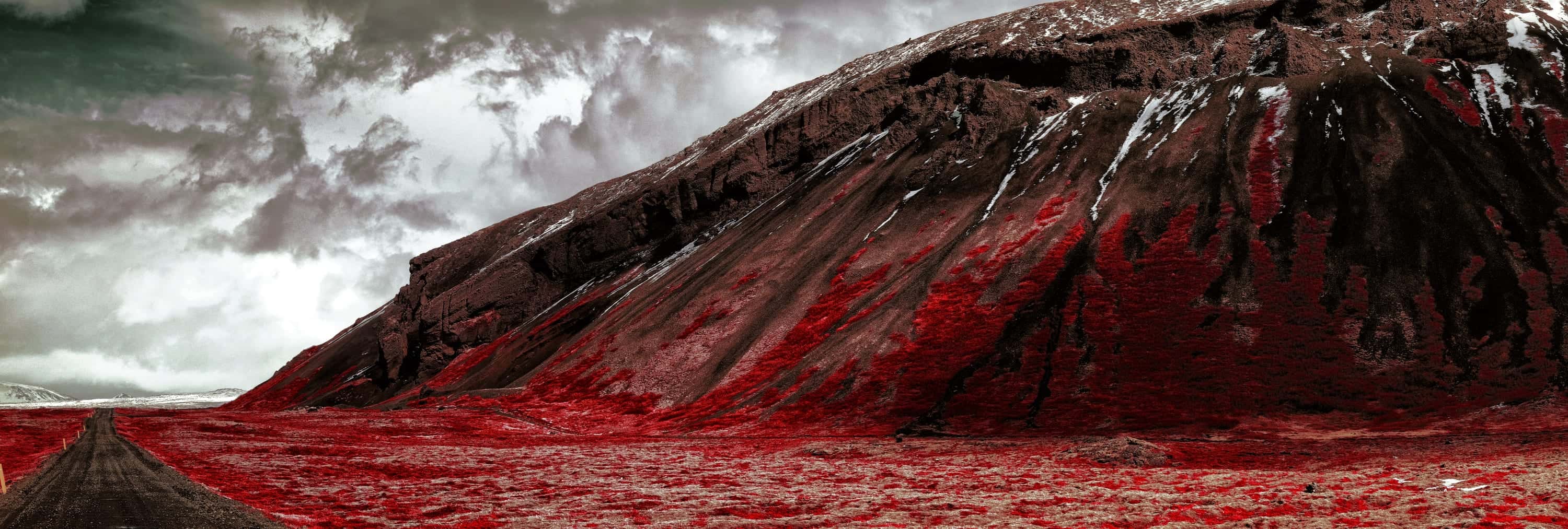 Panorama of a lone road crossing a crimson valley floor beneath a dark snow-streaked massif and storm clouds